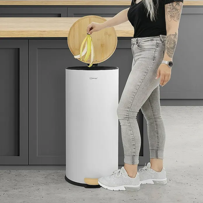 ML-Design Standard Mülleimer Badeimer Abfalleimer 30L rund Weiß Stahl mit Inneneimer Bambusdeckel - 30 l, Stahl, Weiß - Frau wirft Bananenschale in MDesign Treteimer mit Bambusdeckel. Der Eimer ist weiß, und die Küche ist modern mit grauen Schränken.