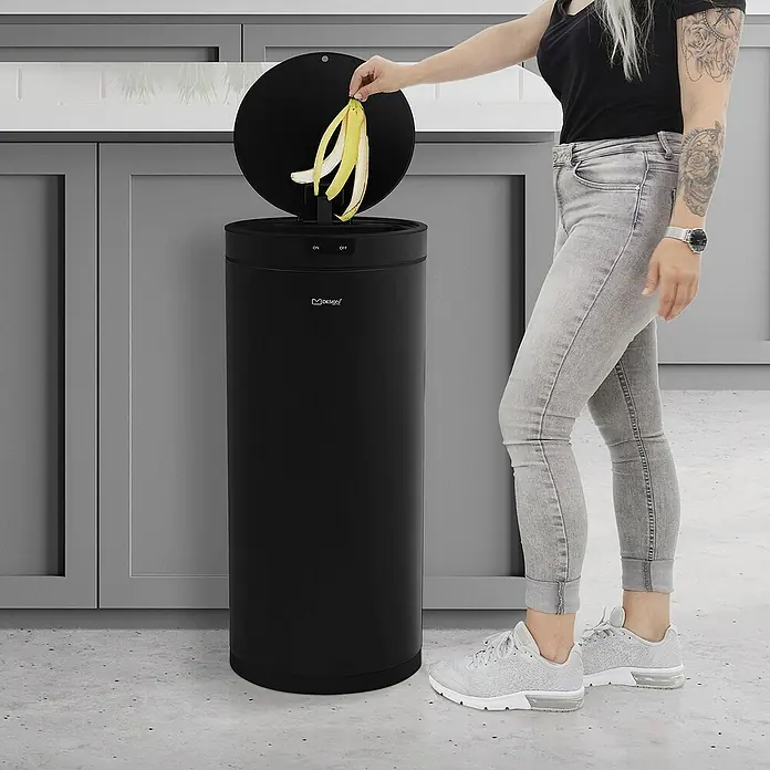ML-Design Sensormülleimer 40L rund aus Stahl Abfalleimer berührungslos, Klemmring für Müllbeutel - 40 l, Stahl, Schwarz - Ein schwarzer, runder Automatik-Mülleimer wird von einer Person mit Bananenschalen befüllt. Der Mülleimer steht in einer modernen Küche.