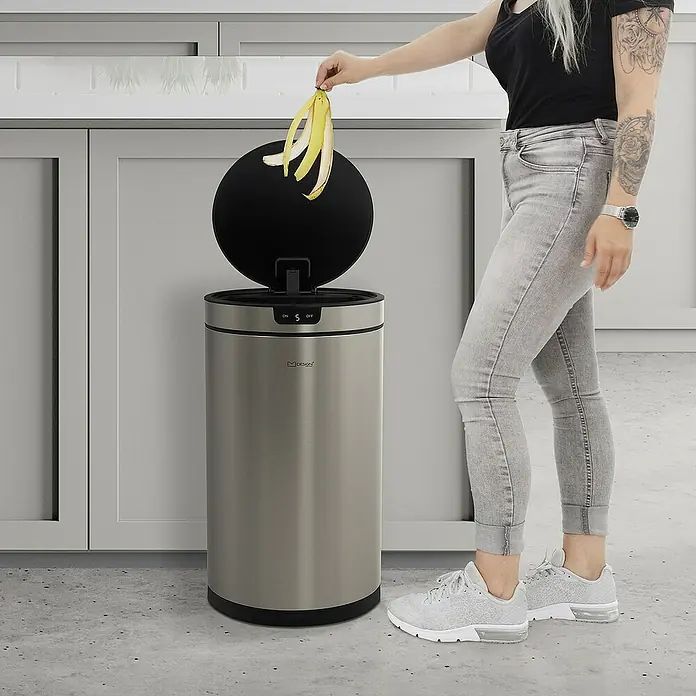 ML-Design Sensormülleimer 30L rund Edelstahl Abfalleimer berührungslos, Klemmring für Müllbeutel - 30 l, Stahl, Silber - Frau wirft Bananenschale in einen MDesign Automatik-Mülleimer aus Edelstahl.