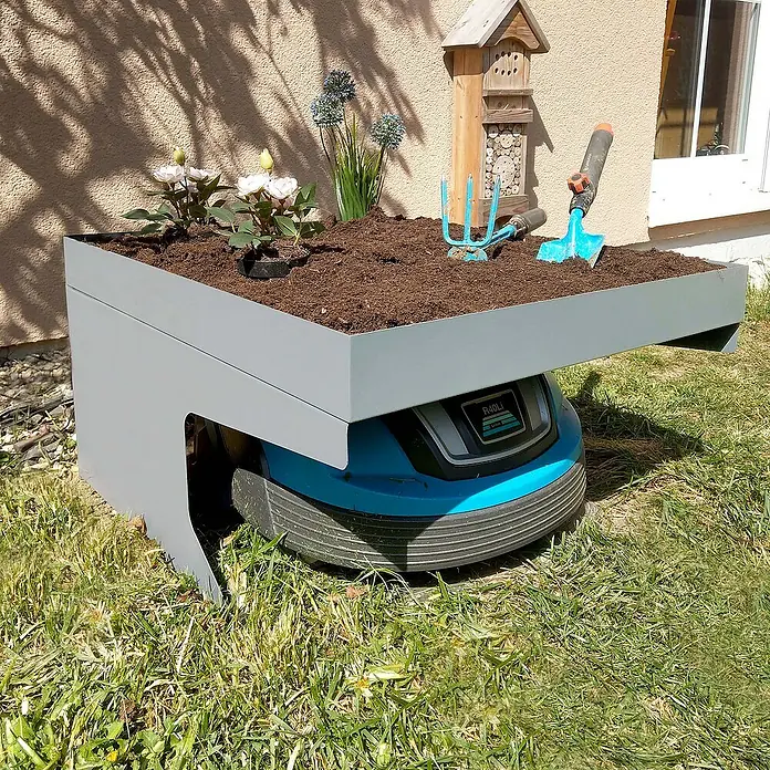 Proregal Mähroboter-Garage Mähroboter-Garage mit Pflanzkasten - Grau - Kreative Ladestation: Ein Rasenmähroboter dient als Basis für einen erhöhten Pflanzkasten mit Blumen und Gartengeräten.