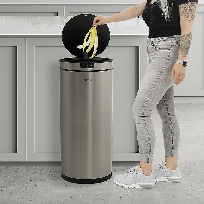 ML-Design Sensormülleimer 40L rund Edelstahl Abfalleimer berührungslos, Klemmring für Müllbeutel - 40 l, Stahl, Silber - Eine Frau wirft Bananenschalen in einen Motion Sensor Mülleimer aus Edelstahl. Der Deckel des Mülleimers ist geöffnet.