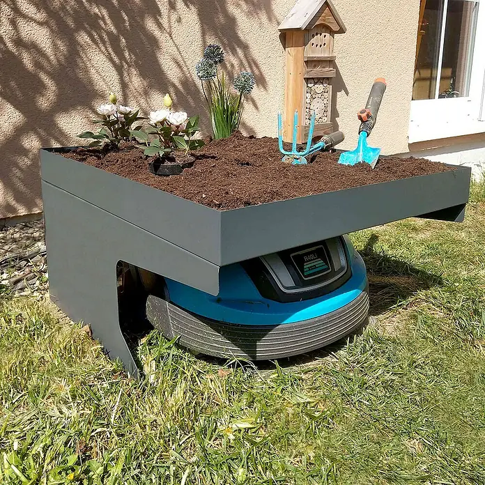 Proregal Mähroboter-Garage Mähroboter-Garage mit Pflanzkasten - Anthrazit - Ein Gardena Rasenmähroboter mit einem aufgesetzten Pflanzkasten, bepflanzt mit Blumen und Gartengeräten, steht auf einer grünen Rasenfläche.