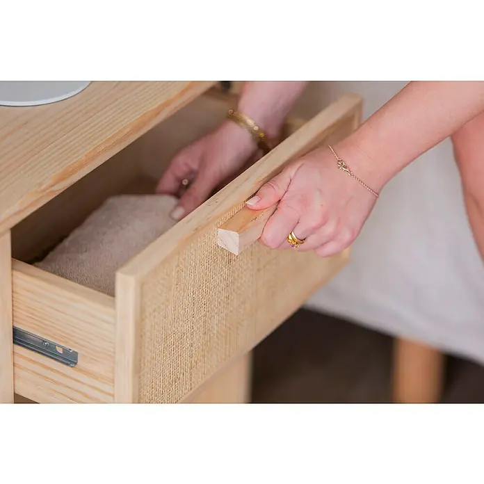 Astigarraga Aura Conjunto de muebles - 5 pzs., Madera maciza y rafia, Natural - Primer plano de una mesita de noche de madera clara con un cajón abierto por una mujer.