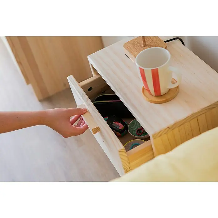 Astigarraga Vita Conjunto de muebles - Natural, 4 pzs. - Una mano abre el cajón de una mesita de noche de madera clara, con una taza encima y varios objetos visibles dentro del cajón.