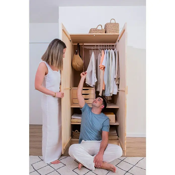 Astigarraga Vita Conjunto de muebles - Natural, 4 pzs. - Una pareja mira un armario de madera abierto lleno de ropa, bolsos y cestas. El mueble tiene un aspecto natural.