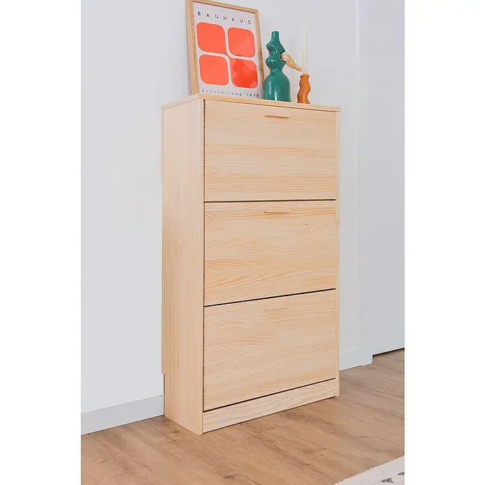 Astigarraga Vita Conjunto de muebles - Natural, 2 pzs. - Un mueble zapatero de madera con tres compartimentos se apoya contra una pared, adornado con decoraciones y un póster de la Bauhaus.
