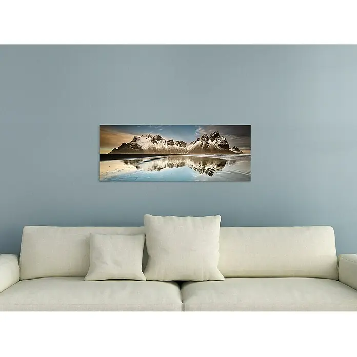 Glasbild - Reflections, B x H: 125 x 50 cm - Leinwandbild mit einer malerischen Berglandschaft, die sich im Wasser spiegelt, über einem weißen Sofa platziert.