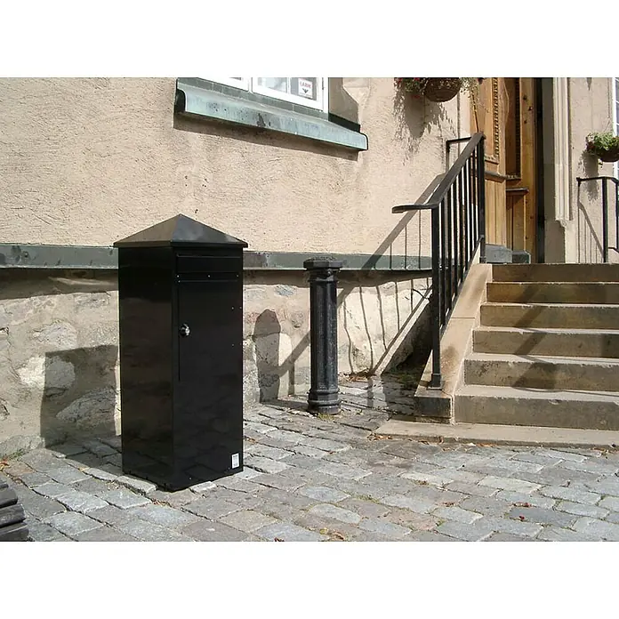 Proregal Wandbriefkasten aus Metal - L x B x H: 39 x 39 x 108 cm, Schwarz, Metall - Der moderne, schwarze Briefkasten mit Pyramidenhut steht auf einem gepflasterten Platz vor einem Gebäude und bietet eine stilvolle Möglichkeit Post zu empfangen.