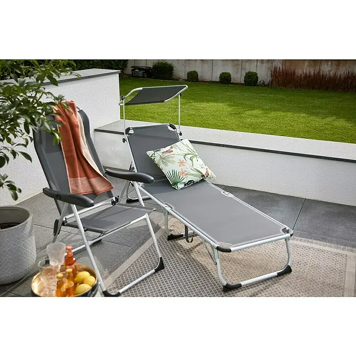 Sunfun Campingliege mit Sonnendach - Night-Grey, Aluminium, Textilene - Ein grauer Liegestuhl mit Sonnendach und Kissen auf einem Teppich, daneben ein passender Stuhl mit Handtuch und Getränken auf einem Tablett.