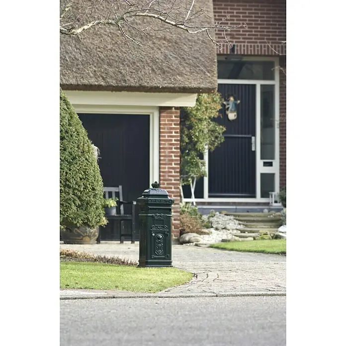 Proregal Standbriefkasten aus Gießaluminium - L x B x H: 36,5 x 33 x 110 cm, Grün, Metall - Diese Aufnahme zeigt einen grünen Briefkasten vor einem Backsteinhaus mit einem strohgedeckten Dach, der eine charmante und einladende Atmosphäre ausstrahlt.