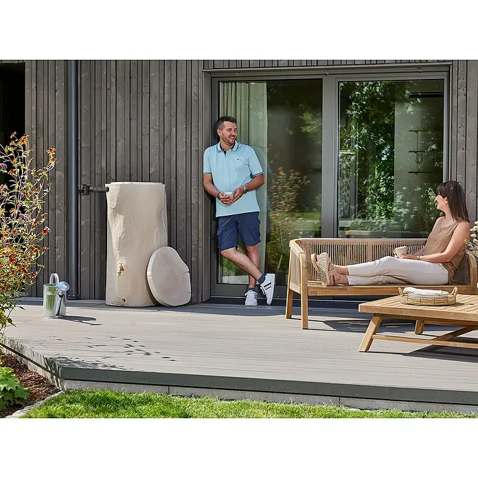 Garantia Regenspeicher - 250 l, Greywood - Das Foto zeigt einen stilvollen Wasserspeicher in Baumstammoptik, der sich perfekt in den Garten einfügt. Er ist neben einer Hauswand mit Holzbeschlag platziert.