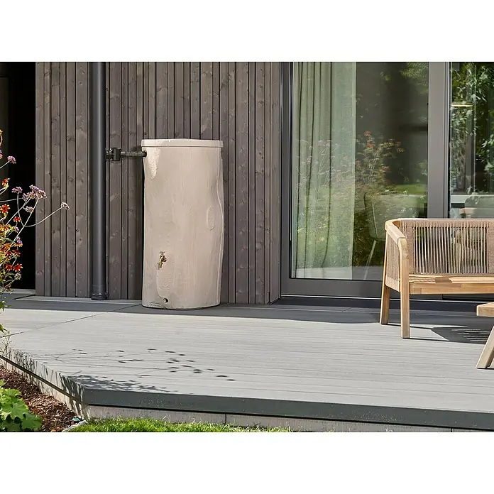 Garantia Regenspeicher - 250 l, Greywood - Nahaufnahme einer hellen Wasserspeicher-Tonne mit Messinghahn, die an einer Hauswand mit Rohrleitung auf einer Terrasse steht.