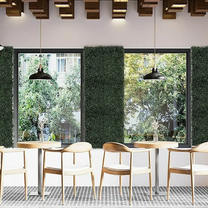 Floordirekt Kunstpflanze Pflanzenwand Boxwood Dark-Green 50 x 50 cm - Blütenfarbe: Grün, Kunststoff - Helle Holzstühle mit weißer Polsterung stehen an Tischen vor Fenstern in einem Café. Die Beleuchtung wird durch moderne Pendelleuchten gewährleistet.