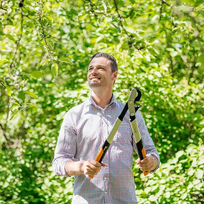 Fiskars Getriebe-Astschere PowerGearX LX94-M - Max. Aststärke: 50 mm, 64 cm - Mann mit einer Fiskars Astschere vor dem Hintergrund von Grünpflanzen, ideal für Gartenarbeit und Baumpflege.