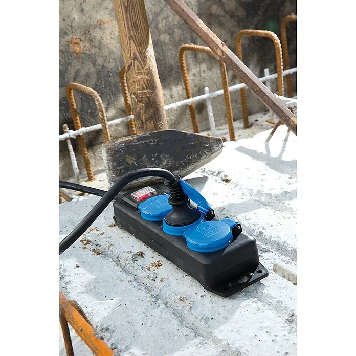 as-schwabe Steckdosenleiste - 3-fach, Schwarz, Kabellänge: 1,5 m, IP44, Mit Schalter - Baustellen-Mehrfachsteckdose mit blauen Schutzkappen und einem eingesteckten Stecker auf einer Baustelle.