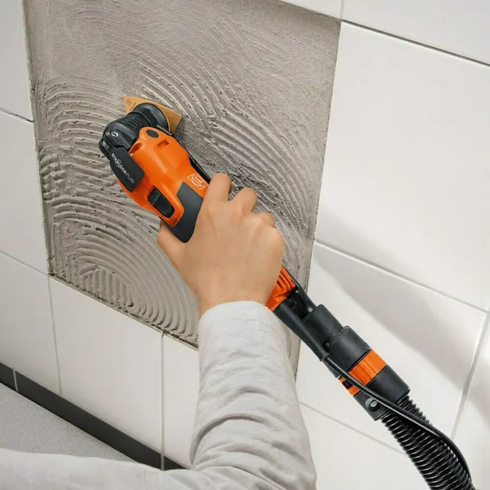 Fein Starlock Hartmetall-Raspel - Breite: 80 mm - Eine Person verwendet ein FEIN Oszilliergerät, um Fliesenkleber von einer Wand zu entfernen. Das Werkzeug ist orange und schwarz.
