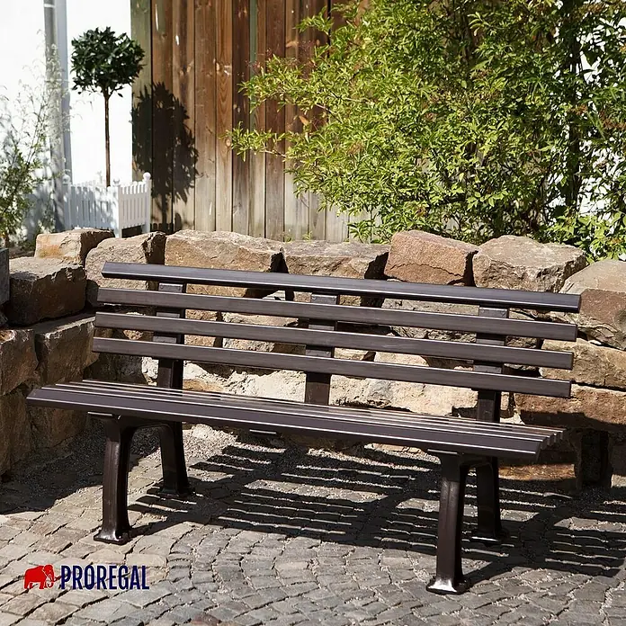 Proregal Gartenbank Antigua 3-Sitzer aus Kunststoff - Braun - Eine elegante Parkbank aus Metall mit dunkler Oberfläche vor einer Steinmauer und Grünpflanzen. Ideal für Gärten und Parks.