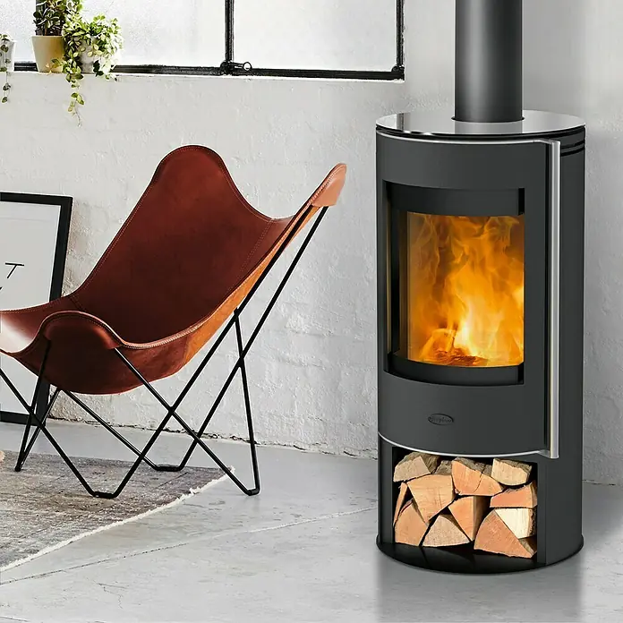 Fireplace Kaminofen Verona - 5 kW, Raumheizvermögen: 90 m³, Material Verkleidung: Stahl, Schwarz - Ein stilvoller Kaminofen mit Holzfach und einem gemütlichen Sessel in einem modernen Wohnraum.