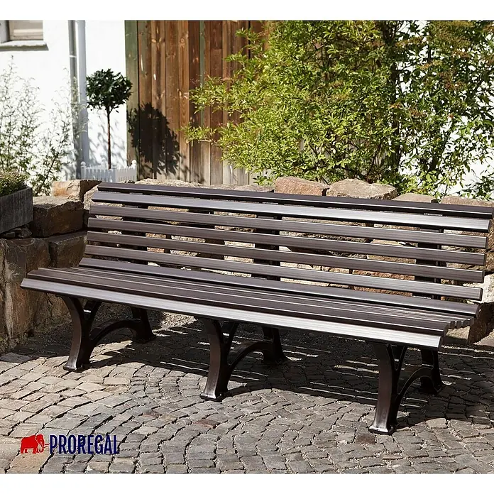 Proregal Gartenbank Jamaika 4-Sitzer aus Kunststoff - Braun - Eine braune Gartenbank mit Lattenrost und geschwungenen Beinen vor einer Steinmauer und einer Holzwand.