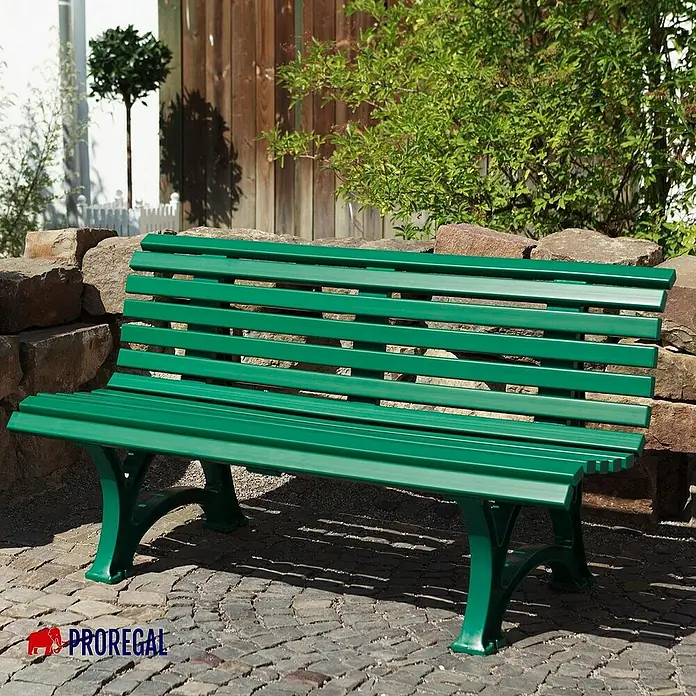 Proregal Gartenbank Jamaika 3-Sitzer aus Kunststoff - Grün - Eine grüne Parkbank aus Holzlatten mit gusseisernen Füßen, die vor einer Steinmauer und grünen Pflanzen steht.