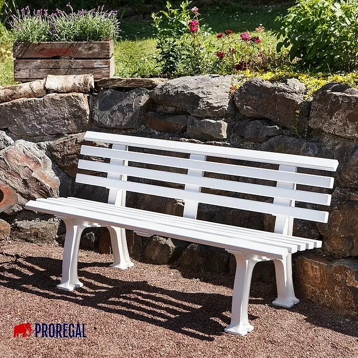 Proregal Gartenbank Antigua 3-Sitzer aus Kunststoff - Weiß - Weiße Parkbank aus Metall mit Lattenmuster vor einer Steinmauer und grünen Pflanzen.