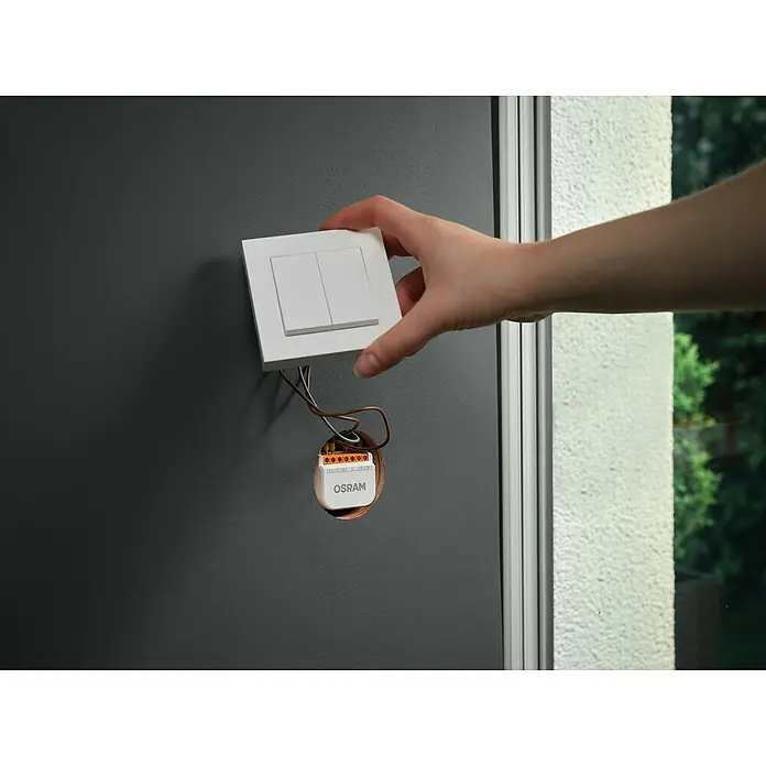 Osram Smart+ Schalter 2CH - Weiß, Innen - Eine Hand montiert einen Osram Smart Home Lichtschalter mit zwei Kippschaltern an einer grauen Wand neben einem Fenster.