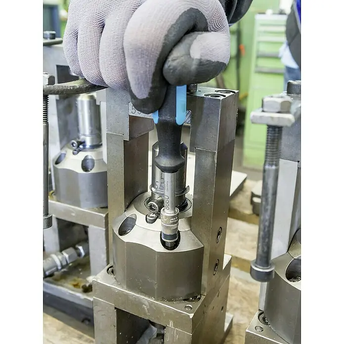 Hazet Steckschlüsseleinsatz-Set 853-1 - ¼″, 36 -tlg. - Ein Arbeiter mit Handschuhen justiert ein silbernes Werkzeug mit blauem Griff in einer Metallvorrichtung in einer Werkstatt.