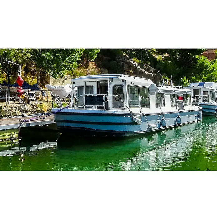 Sunset Solarmodul-Set Laminat - Nennleistung: 30 W, Für Booten, Yachten und Wohnmobilen - Dieses Bild zeigt ein weißes und blaues Hausboot, das friedlich an einem grünen See liegt. Das Boot ist mit einem Steg verbunden.