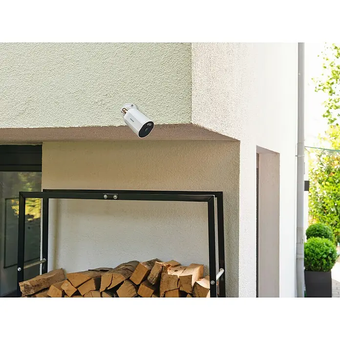 Osram Smart+ Überwachungskamera Outdoor Fixed - Passend für: Alarmsystem, Smart Home System - Bild einer weißen Überwachungskamera an einer Hauswand mit Holzstapel im Vordergrund.