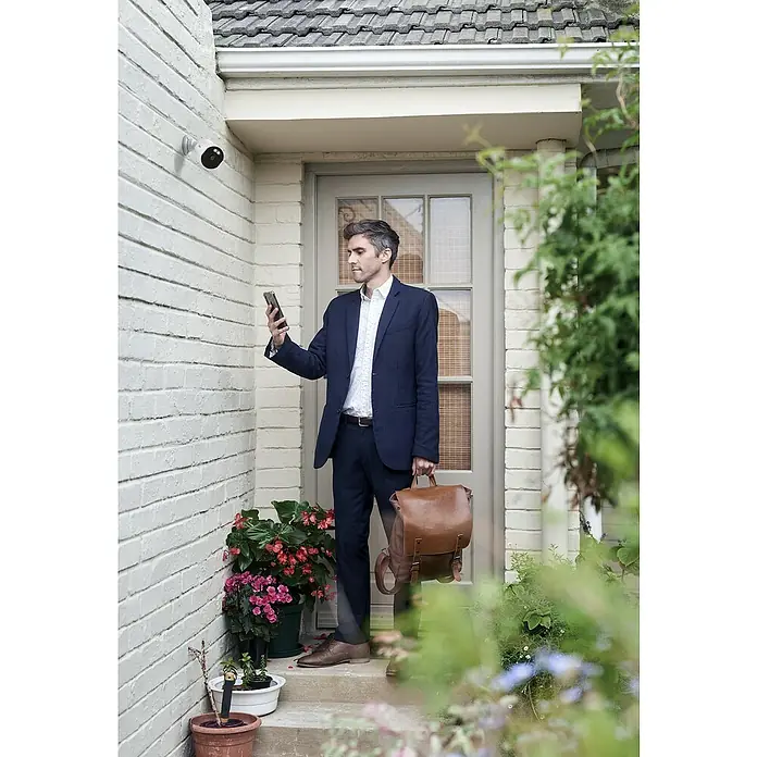 Osram Smart+ Überwachungskamera Outdoor Fixed - Passend für: Alarmsystem, Smart Home System - Ein Mann in einem Anzug steht vor einer Haustür und bedient ein Smart Home Sicherheitssystem mit einer Kamera.
