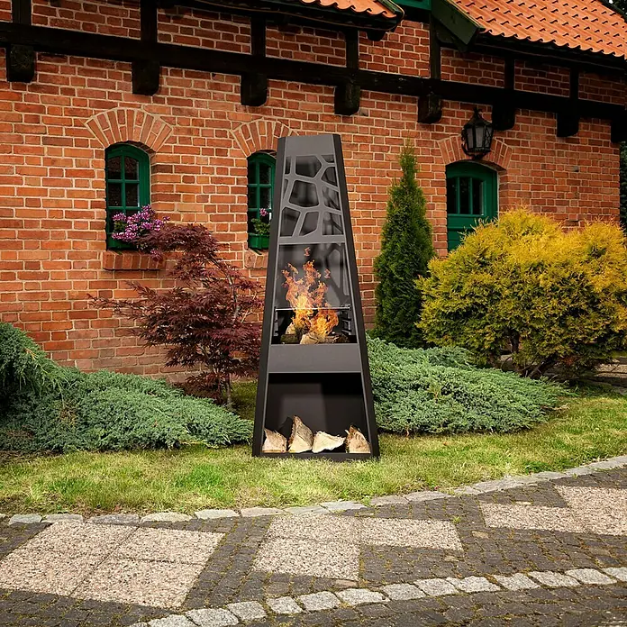 Dobar Gartenkamin - 57,5 x 38 x 148 cm, Stahl, Pulverbeschichtet - Moderner schwarzer Kamin im Freien mit Holzscheiten, umgeben von üppigem Grün, vor einem Backsteingebäude für eine warme Atmosphäre.