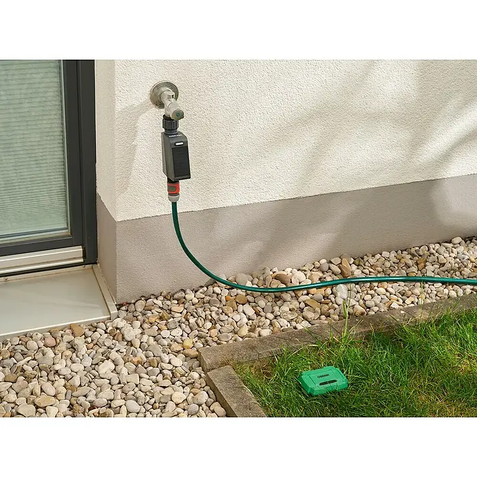 Osram Smart+ Bewässerungssteuerung - Batteriebetrieben, Passend für: Bewässerungssteuerung, Smart Home System, 1 & ¾ Zoll Wasserhahn - Bewässerungssystem: Eine graue Zeitschaltuhr von Osram ist an einem Wasserhahn befestigt und mit einem grünen Schlauch verbunden, der zu einem grünen Sensor führt.