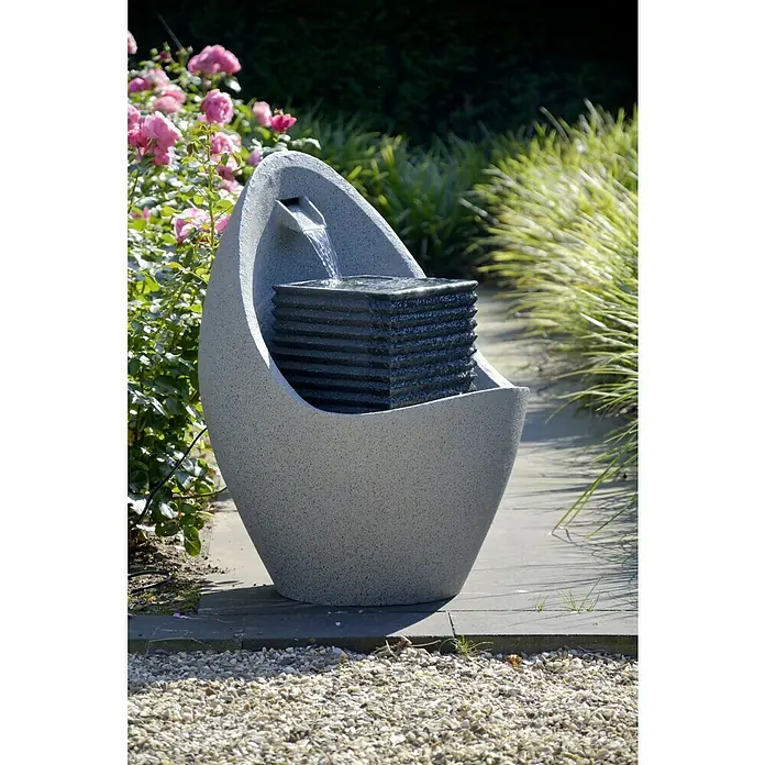 Dobar Kunststeinbrunnen - 49,5 x 49,5 x 66,5 cm - Eine moderne Gartenbrunnen mit grauem Korpus und dunklem Quader, aus dem Wasser fließt, inmitten eines blühenden Gartens.