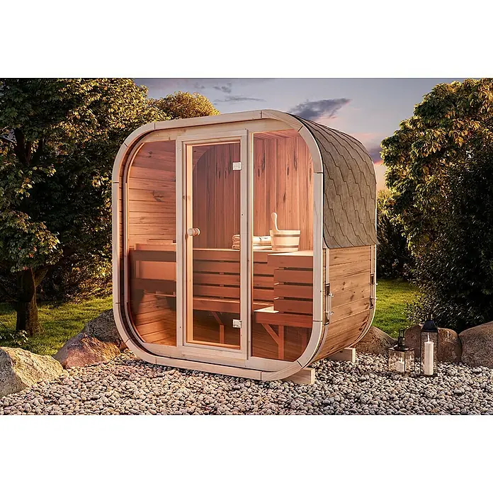 FinnTherm Fasssauna Comfort Set Ellipso M - Elektroofen, Holz - Holzfass-Sauna im Freien mit Glastür und Schindeldach, umgeben von Bäumen und Laternen, die eine entspannende Atmosphäre schaffen.
