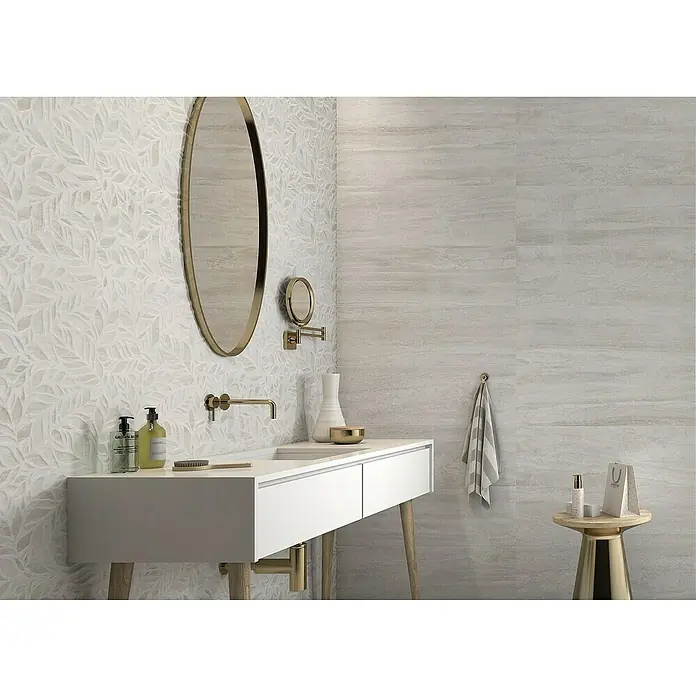 Revestimiento de pared Lumia - 30 x 90 cm, Blanco, Rectificado - Un baño con encimera blanca, accesorios dorados y espejo ovalado, con azulejos de pared claros con estampado de hojas de fondo.