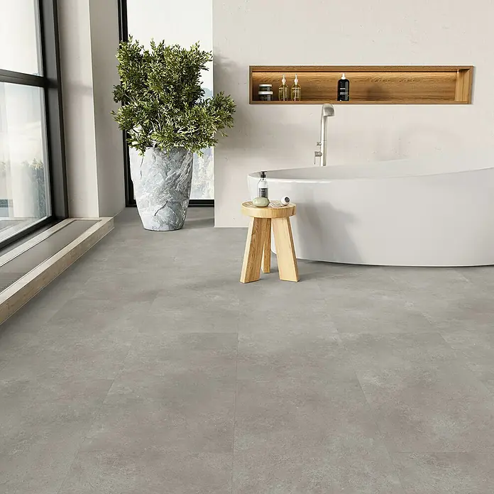 Parador Vinylboden Basic 5.3 - Stein Trafalgar hellgrau, Fliese, 914 x 457 x 5,3 mm - Ein Badezimmer mit grauen Bodenfliesen, einer Badewanne und einem Holzhocker. Das graue Fliesenmuster sorgt für ein elegantes Aussehen.