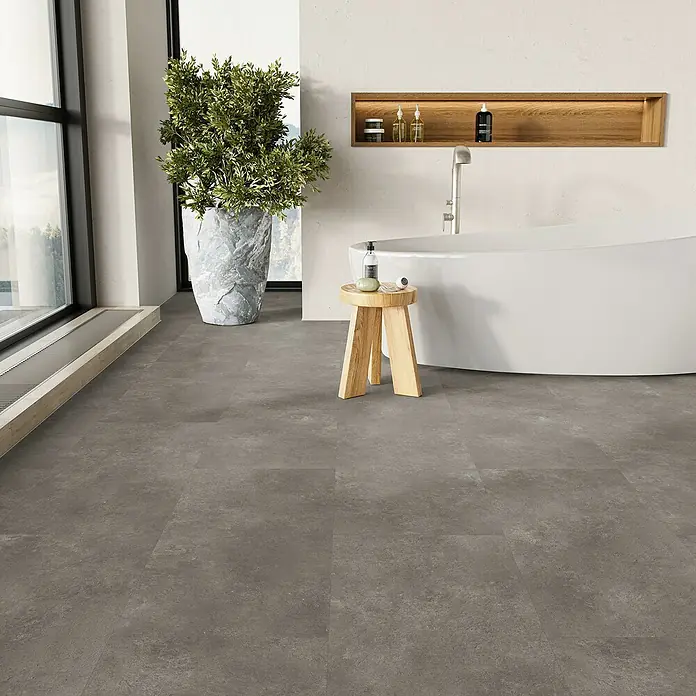 Parador Vinylboden Basic 5.3 - Stein Trafalgar dunkelgrau, Fliese, 914 x 457 x 5,3 mm - Das Bild zeigt einen modernen Badezimmerboden mit grauem Fliesenmuster, ergänzt durch eine hölzerne Ablage, eine grüne Pflanze und eine Badewanne.