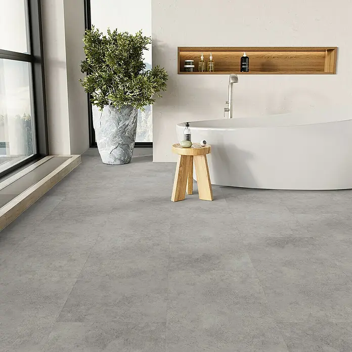 Parador Vinylbodenmuster Basic 5.3 Beton Grau - 200 x 457 x 5,3 mm, Fliesenoptik - Badezimmer mit grauen Bodenfliesen, einer freistehenden Badewanne und einem Holzhocker.