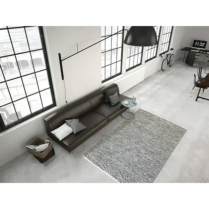 Kayoom Flachgewebeteppich Cocktail 300 - Grau, 150 x 80 cm, 100% Baumwolle - Ein stilvolles Wohnzimmer mit einem modernen schwarzen Sofa, einem gemusterten Teppich und großen Fenstern. Die Einrichtung ist minimalistisch und elegant.
