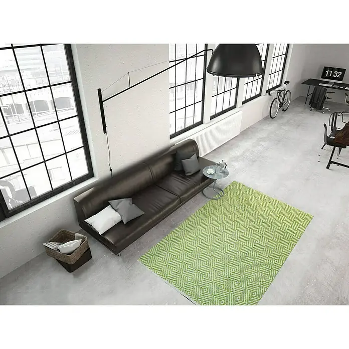 Kayoom Flachgewebeteppich Cocktail 300 - Grün, 150 x 80 cm, 100% Baumwolle - Ein grüner Teppich mit geometrischem Muster liegt in einem modernen Wohnzimmer mit Ledersofa und großen Fenstern.