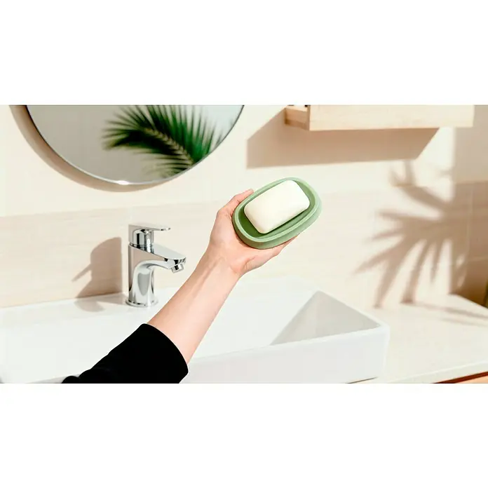 Venus Flakoni Seifenschale - Sage, Polyresin - Eine Hand hält eine grüne Seifenschale mit einem weißen Seifenstück über einem Waschbecken, mit einem Wasserhahn und einem runden Spiegel im Hintergrund.