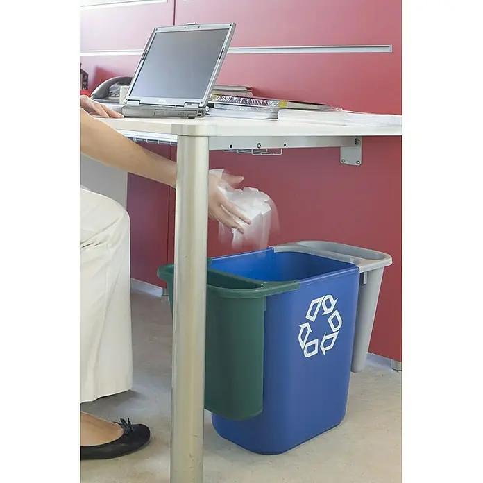 Proregal Standard-Mülleimer Rubbermaid rechteckiger Abfallbehälter aus Polyethylen - 26,6 l, Kunststoff, Blau mit Recyclingsymbol - Ein blauer Recycling-Behälter mit einem grünen Einsatz und dem Recycling-Symbol steht unter einem Schreibtisch, während jemand Abfall hineinwirft.