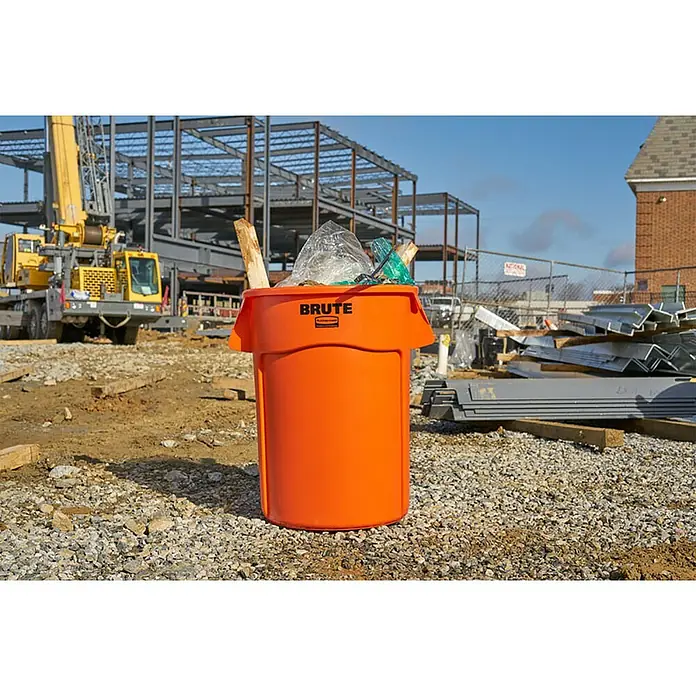 Proregal Standard-Mülleimer Rubbermaid Brute runde Mehrzwecktonne mit Belüftungskanälen - 166,5 l, Kunststoff, Orange - Nahaufnahme eines orangefarbenen, robusten Mülleimers der Marke Rubbermaid auf einer Baustelle, gefüllt mit Abfall, der seine Langlebigkeit hervorhebt.