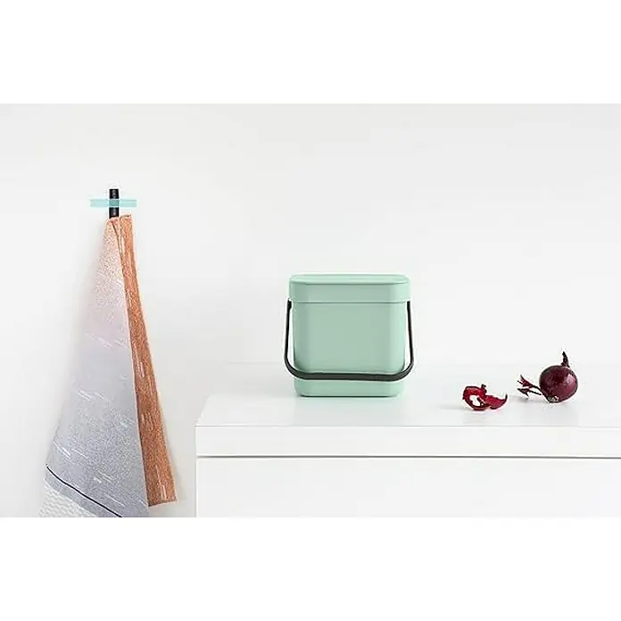 Proregal Standard Mülleimer Brabantia Abfallsammler mit Stay-open Deckel - 3 l, Kunststoff, Jade - Ein mintgrüner Bioeimer mit schwarzem Griff steht auf einem weißen Tisch, daneben eine rote Zwiebel. An der Wand hängt ein Handtuch.