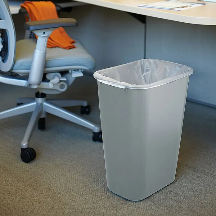 Proregal Standard-Mülleimer Rubbermaid rechteckiger Abfallbehälter - 39 l, Kunststoff, Grau - Ein grauer, eckiger Papierkorb mit einem durchsichtigen Müllbeutel darin steht neben einem Bürostuhl auf einem Teppichboden.