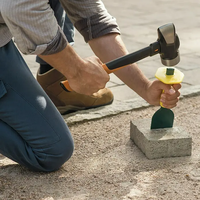 Fiskars Fäustel M - Gewicht Kopf: 1.340 g, Material Stiel: Glasfaserverstärkter Kunststoff (GfK) - Nahaufnahme eines Mannes, der einen Spalthammer und einen Pflastermeißel zum Spalten eines Ziegelsteins auf einer Baustelle verwendet.