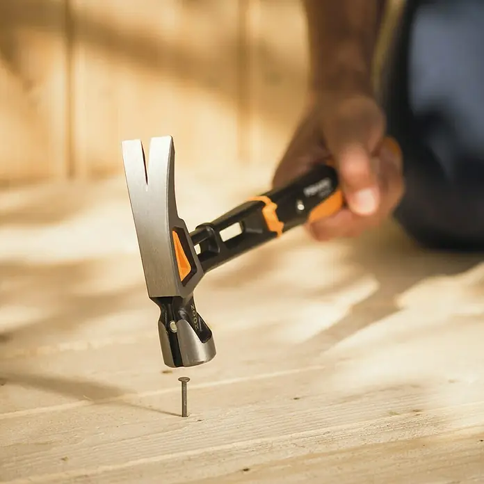 Fiskars Universalhammer XL - Gewicht Kopf: 570 g, Länge: 390 mm - Nahaufnahme eines Fiskars-Hammers, der einen Nagel in ein Holzbrett schlägt, wobei der Fokus auf dem Werkzeug und seiner Funktionalität liegt.