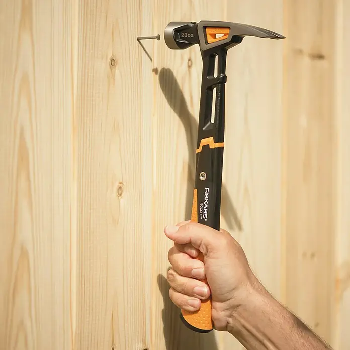 Fiskars Universalhammer L - Gewicht Kopf: 570 g, Länge: 340 mm - Nahaufnahme eines Fiskars IsoCore-Hammers, der einen Nagel in ein Holzbrett treibt. Das Werkzeug ist robust und ergonomisch gestaltet.