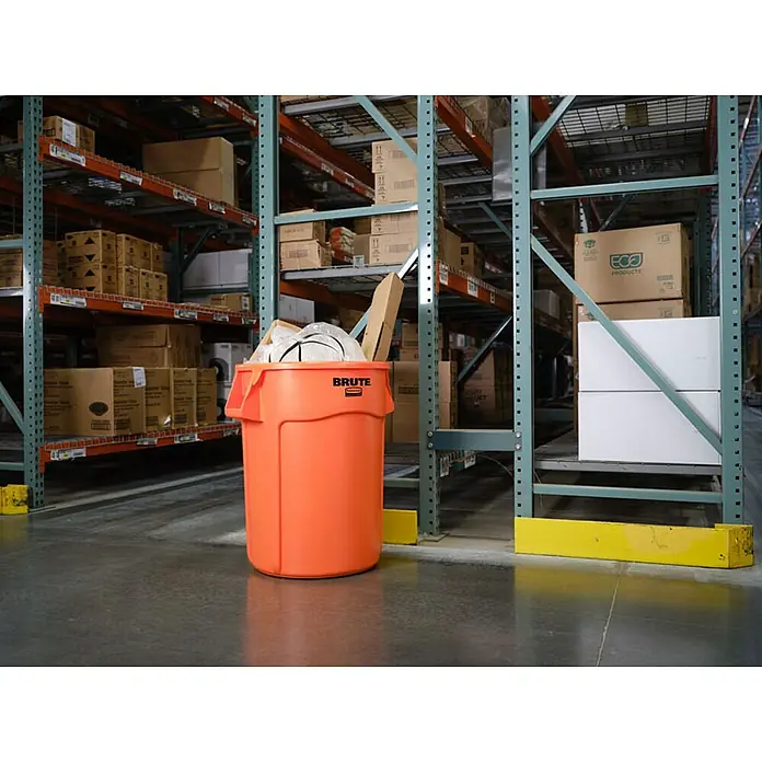 Proregal Standard-Mülleimer Rubbermaid Brute runde Mehrzwecktonne mit Belüftungskanälen - 121,1 l, Kunststoff, Orange - Ein orangefarbener Brute-Mülleimer in einem Lagerhaus, umgeben von Lagerregalen voller Kartons und Waren.