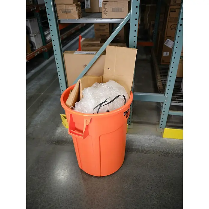 Proregal Standard-Mülleimer Rubbermaid Brute runde Mehrzwecktonne mit Belüftungskanälen - 121,1 l, Kunststoff, Orange - Ein orangefarbener Brute-Abfallbehälter gefüllt mit Kartons und Plastikmüll steht in einem Lagerhaus.